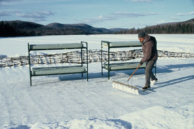 morelli in ice skates sweeps frozen lake with two bunk beds in background